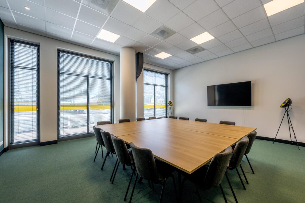 Modern conference room at Elizabeth Cady Stantonplein with a large square meeting table, chairs, flat screen TV, and floor-to-ceiling windows with blinds.